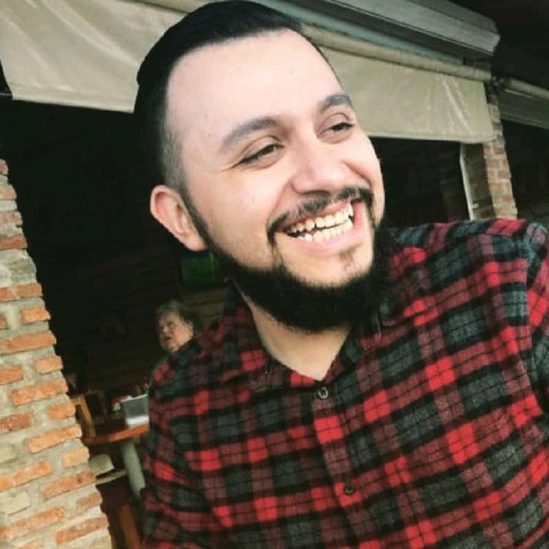 Smiling man with beard in red plaid shirt at outdoor cafe.